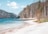 A serene lakeside scene with clear blue water, a small wooden hut beside the lake, and a backdrop of forested mountains and trees. The shore is sandy and curves gently along the water's edge.