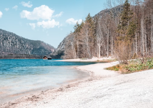 A serene lakeside scene with clear blue water, a small wooden hut beside the lake, and a backdrop of forested mountains and trees. The shore is sandy and curves gently along the water's edge.