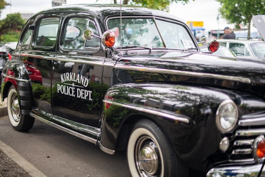 A vintage black police car with chrome accents is parked on a street. The side of the car displays the words 'Kirkland Police Dept.' The car features a reflective surface and classic design elements, including white-walled tires and red signal lights mounted on the roof.