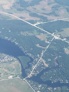 An aerial view of a landscape featuring a network of roads intersecting over a patchwork of greenery and waterways. A river or large body of water is visible, with several smaller streams branching out. There are clusters of buildings and fields, as well as dense areas of trees and vegetation.