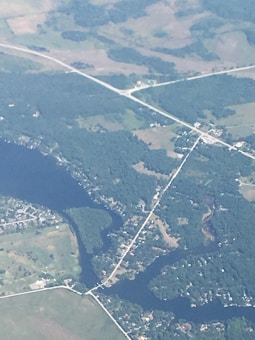 An aerial view of a landscape featuring a network of roads intersecting over a patchwork of greenery and waterways. A river or large body of water is visible, with several smaller streams branching out. There are clusters of buildings and fields, as well as dense areas of trees and vegetation.