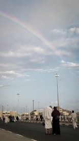 A street scene with a large crowd of people dressed in robes, possibly participating in a pilgrimage. Overhead, a faint rainbow arcs across a mostly cloudy sky. The background features buildings and streetlights.