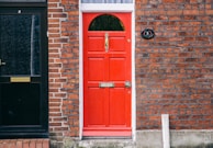 A bright red door with a brass mail slot and door knocker is set against a brick wall. The door displays the number 5 and a small sign stating 'NO JUNK MAIL.' Adjacent to it is a black door with intricate patterns and a mail slot. The surrounding brickwork has a mix of red and dark hues.