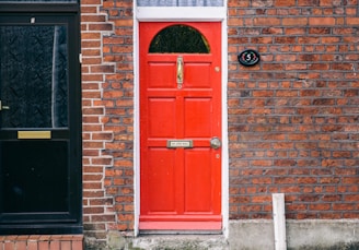 A bright red door with a brass mail slot and door knocker is set against a brick wall. The door displays the number 5 and a small sign stating 'NO JUNK MAIL.' Adjacent to it is a black door with intricate patterns and a mail slot. The surrounding brickwork has a mix of red and dark hues.