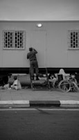 A person is standing on a set of steps leading up to a door on a modular building. The building has barred windows on either side of the door. Nearby on the pavement, there is a wheelchair and another person sitting on the ground. The overall scene is in black and white.