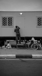 A person is standing on a set of steps leading up to a door on a modular building. The building has barred windows on either side of the door. Nearby on the pavement, there is a wheelchair and another person sitting on the ground. The overall scene is in black and white.