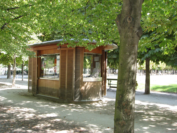 A wooden kiosk nestled among lush green trees beside a sparkling community pool on a sunny summer day.