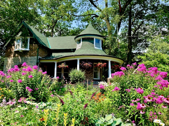 A charming stone cottage with a green roof is surrounded by a lush garden filled with vibrant pink and purple flowers. Tall trees frame the scene, casting dappled sunlight across the landscape. The cottage features a rounded porch with hanging baskets of red flowers, adding to the quaint and inviting appearance.