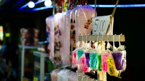 A variety of colorful items are displayed in plastic bags, hanging alongside packaged cotton candy. Each item is priced at 5000 as indicated by a sign. The scene is set in a bright, likely festive environment with various lights and decorations visible in the background.