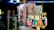 A variety of colorful items are displayed in plastic bags, hanging alongside packaged cotton candy. Each item is priced at 5000 as indicated by a sign. The scene is set in a bright, likely festive environment with various lights and decorations visible in the background.