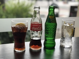 A wooden table displays two bottles and two glasses of soda: a glass with Coca-Cola containing ice and lemon, an empty Coca-Cola bottle, a Sprite bottle, and a glass of Sprite with ice and lemon. A blurred outdoor scene provides the background.