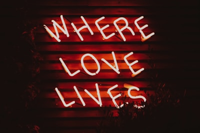 Neon letters spelling out 'Where Love Lives' are displayed on a dark wooden background. The glowing letters create a warm, inviting atmosphere.
