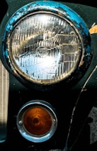A close-up view of a vintage car's headlight and turn signal. The large circular headlight is framed by a blue, weathered rim showing signs of rust and chipping paint. Below it, a smaller orange signal light with concentric circles is encased in a silver ring. Both lights are attached to a dark green car body.
