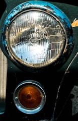 A close-up view of a vintage car's headlight and turn signal. The large circular headlight is framed by a blue, weathered rim showing signs of rust and chipping paint. Below it, a smaller orange signal light with concentric circles is encased in a silver ring. Both lights are attached to a dark green car body.