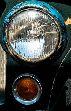 A close-up view of a vintage car's headlight and turn signal. The large circular headlight is framed by a blue, weathered rim showing signs of rust and chipping paint. Below it, a smaller orange signal light with concentric circles is encased in a silver ring. Both lights are attached to a dark green car body.