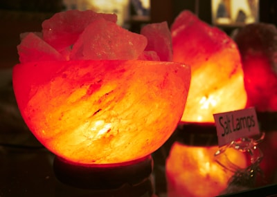 A glowing salt lamp, crafted from pink Himalayan salt, emits a warm and soothing orange light. The lamp is placed on a reflective surface, enhancing its luminous glow. In the background, another salt lamp adds to the ambient lighting. A small sign labeled 'Salt Lamps' is visible, indicating the type of object.
