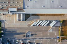 An aerial view of a large distribution center or warehouse with several loading docks and parked trucks. The area is organized with clear lines and parking spaces. The roof of the warehouse is visible, highlighting its industrial design. There are a few cars in the parking area, indicating activity but not congestion.