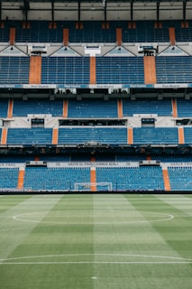 A large, empty football stadium with rows of blue and orange seats. The center of the image focuses on a green football pitch with neatly maintained grass, and the goalposts are visible in the distance. The stadium is adorned with signage and banners, indicating it is associated with Real Madrid.