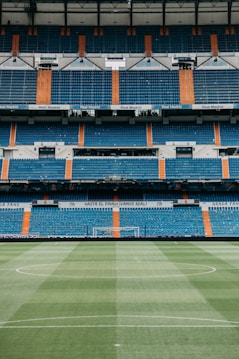 A large, empty football stadium with rows of blue and orange seats. The center of the image focuses on a green football pitch with neatly maintained grass, and the goalposts are visible in the distance. The stadium is adorned with signage and banners, indicating it is associated with Real Madrid.