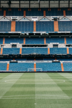A large, empty football stadium with rows of blue and orange seats. The center of the image focuses on a green football pitch with neatly maintained grass, and the goalposts are visible in the distance. The stadium is adorned with signage and banners, indicating it is associated with Real Madrid.
