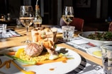 A well-prepared dining table featuring a gourmet dish with a piece of meat served on a bed of yellow grains, garnished with asparagus and a sauce swirl. There are two glasses of white wine, a bottle of olive oil, a basket of sliced bread, and a side salad in a bowl. The setting is warmly lit, creating a cozy and inviting atmosphere.