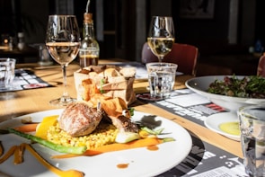 A well-prepared dining table featuring a gourmet dish with a piece of meat served on a bed of yellow grains, garnished with asparagus and a sauce swirl. There are two glasses of white wine, a bottle of olive oil, a basket of sliced bread, and a side salad in a bowl. The setting is warmly lit, creating a cozy and inviting atmosphere.
