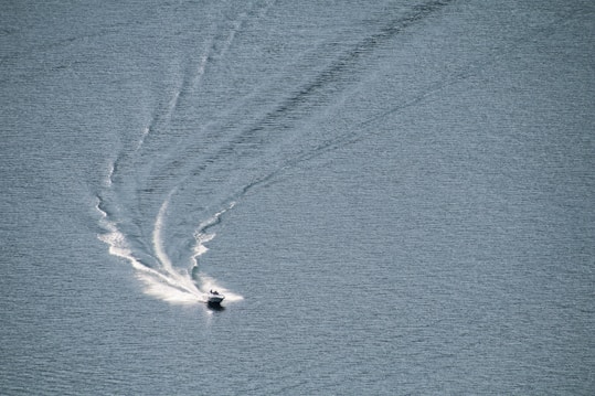 A small motorboat moves across a vast body of water, leaving a wide wake behind it. The surface of the water is rippled and has a muted blue-gray tone, with the boat creating dynamic white trails as it speeds forward.