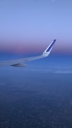 An airplane wing is visible against a backdrop of a serene sky transitioning from a deep blue to a soft pink hue. The ground is faintly visible far below, indicating the plane is at a high altitude. The logo 'SAS' is visible on the wing tip.