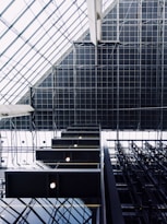 Geometric patterns and architectural elements dominate the visual space, with a focus on the modern interior of a building. Large glass panels create a grid-like pattern on a ceiling, while rectangular light fixtures add a symmetrical contrast. Industrial materials such as steel beams contribute to the modern aesthetic.