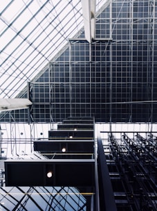 Geometric patterns and architectural elements dominate the visual space, with a focus on the modern interior of a building. Large glass panels create a grid-like pattern on a ceiling, while rectangular light fixtures add a symmetrical contrast. Industrial materials such as steel beams contribute to the modern aesthetic.