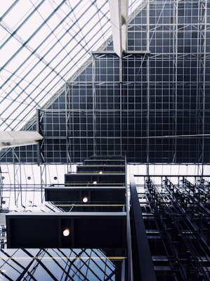 Geometric patterns and architectural elements dominate the visual space, with a focus on the modern interior of a building. Large glass panels create a grid-like pattern on a ceiling, while rectangular light fixtures add a symmetrical contrast. Industrial materials such as steel beams contribute to the modern aesthetic.