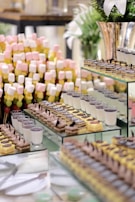 An arrangement of various desserts on display, including layers of cakes, panna cotta, and skewers of marshmallows in pastel colors. The presentation is on glass shelves with additional decorative elements like plants in the background.