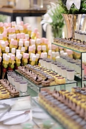 An arrangement of various desserts on display, including layers of cakes, panna cotta, and skewers of marshmallows in pastel colors. The presentation is on glass shelves with additional decorative elements like plants in the background.