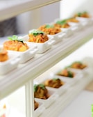 A series of small, white, square dishes arranged in rows on wooden shelves, each containing a portion of appetizing, spiced food garnished with cilantro.
