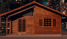 A small wooden house with a rustic appearance, featuring a traditional gabled roof and closed wooden shutters on the window. The facade is made of horizontal wooden planks and there is a set of double doors with glass panes. The house is surrounded by some greenery and trees, with part of a sign visible on the right side.