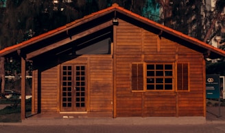 A small wooden house with a rustic appearance, featuring a traditional gabled roof and closed wooden shutters on the window. The facade is made of horizontal wooden planks and there is a set of double doors with glass panes. The house is surrounded by some greenery and trees, with part of a sign visible on the right side.