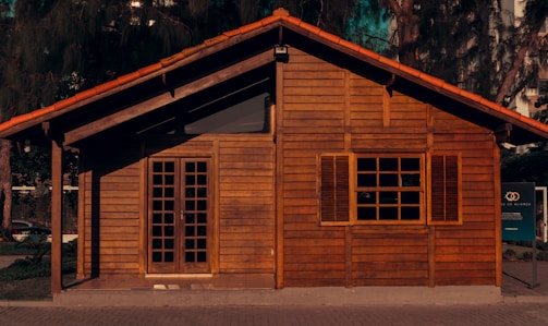 A small wooden house with a rustic appearance, featuring a traditional gabled roof and closed wooden shutters on the window. The facade is made of horizontal wooden planks and there is a set of double doors with glass panes. The house is surrounded by some greenery and trees, with part of a sign visible on the right side.