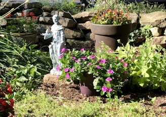 A serene garden scene featuring a small statue of a figure dressed in robes, surrounded by lush green plants and vibrant flowers in bloom. The setting includes a stone wall in the background, adding a rustic touch to the environment. The sunlight casts gentle shadows, enhancing the natural beauty of the scene.