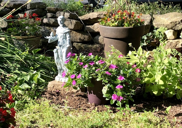 A serene garden scene featuring a small statue of a figure dressed in robes, surrounded by lush green plants and vibrant flowers in bloom. The setting includes a stone wall in the background, adding a rustic touch to the environment. The sunlight casts gentle shadows, enhancing the natural beauty of the scene.