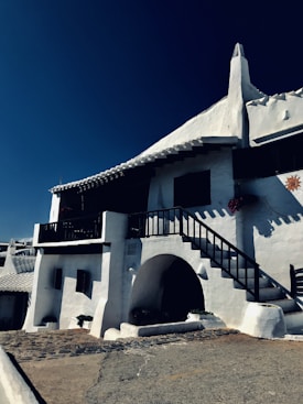 A white stucco building with dark wood railings and a staircase leading to an upper level. The architecture features curved lines and an archway. Bright sunlight casts shadows, and there are decorative elements like plants and a sun motif on the wall.