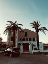 A large, Mediterranean-style villa with a warm-colored facade, surrounded by tall palm trees. The house has symmetrical features, with black shuttered windows and a grand entrance with steps leading up to a balcony. Below, a dark SUV is parked near the entrance, and the sky is clear and blue.