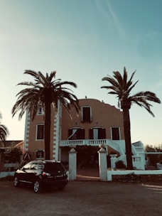 A large, Mediterranean-style villa with a warm-colored facade, surrounded by tall palm trees. The house has symmetrical features, with black shuttered windows and a grand entrance with steps leading up to a balcony. Below, a dark SUV is parked near the entrance, and the sky is clear and blue.