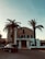 A large, Mediterranean-style villa with a warm-colored facade, surrounded by tall palm trees. The house has symmetrical features, with black shuttered windows and a grand entrance with steps leading up to a balcony. Below, a dark SUV is parked near the entrance, and the sky is clear and blue.