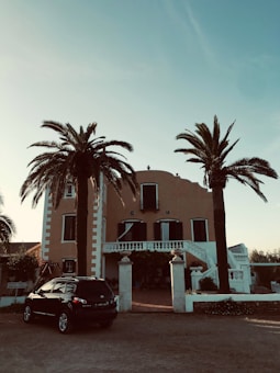 A large, Mediterranean-style villa with a warm-colored facade, surrounded by tall palm trees. The house has symmetrical features, with black shuttered windows and a grand entrance with steps leading up to a balcony. Below, a dark SUV is parked near the entrance, and the sky is clear and blue.