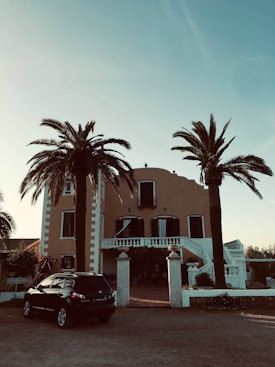 A large, Mediterranean-style villa with a warm-colored facade, surrounded by tall palm trees. The house has symmetrical features, with black shuttered windows and a grand entrance with steps leading up to a balcony. Below, a dark SUV is parked near the entrance, and the sky is clear and blue.