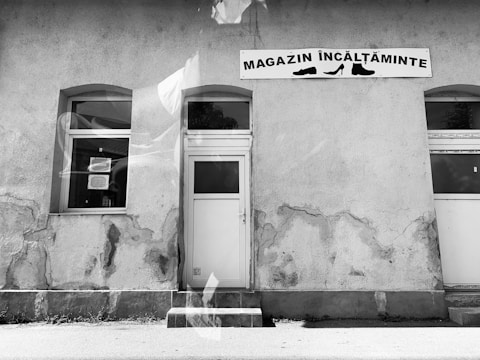 A weathered storefront wall with a sign reading 'Magazin Încălțăminte', featuring shoe graphics. The building facade shows signs of age, with cracked and peeling plaster. A white door and a window are positioned along the wall, creating a symmetrical appearance. The monochrome effect accentuates the textures and contrasts in the scene.