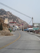 A paved road leads through a mountainous area, lined by hills with rocky outcrops. There are multiple buildings, including a mix of residential and commercial properties. Vehicles are parked along the roadside, and utility poles with wires stretch across the scene. A sign with Chinese characters is visible, and the landscape has a slightly overcast sky.