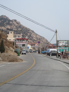 A paved road leads through a mountainous area, lined by hills with rocky outcrops. There are multiple buildings, including a mix of residential and commercial properties. Vehicles are parked along the roadside, and utility poles with wires stretch across the scene. A sign with Chinese characters is visible, and the landscape has a slightly overcast sky.