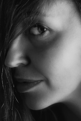 A close-up, monochromatic photograph featuring the side profile of a person's face. The image focuses on the eye, nose, lips, and part of the hair, with detailed skin texture and soft shadows enhancing the facial features.