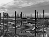 A construction site with an unfinished steel framework structure, surrounded by trees and a cloudy sky. Various construction materials and vehicles are visible at ground level within the site.